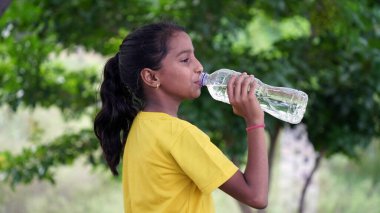 Parkta siyah saçlı, su içen, plastik şişe tutan ve baş parmağını kaldıran mutlu Hintli kız. Açık hava çekimi