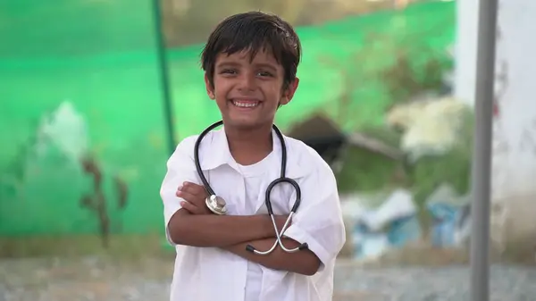 Küçük şirin Hintli çocuk elinde steteskop, elinde doktorculuk oynayan doktor üniforması, kameraya bakan çocuk doktoru rolü yapan neşeli, küçük, karışık ırk anaokulu çocuğu.