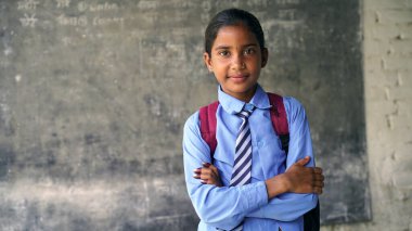 Sırt çantalı, kameraya bakan gülümseyen Hintli kırsal okul çocuğu. Gülümseyen bir okul üniforması giyen neşeli bir çocuk. İlkokul ve ilköğretim.
