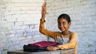 Mutlu Hintli okul çocuğunun portresi sınıfta kitap ve dizüstü bilgisayarla çalışırken el kaldırıyor. Çevrimiçi ders öğreniliyor.