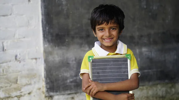 Portrait of happy cute little indian boy in school uniform holding blank slate, Adorable elementary kid showing black board. child education concept. rural india.
