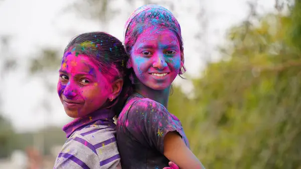 Happy Indian friends or kids celebrating the Hindu festival of Holi by applying colors to each other, best friends. Female siblings holding a plate with different shades of Gulal