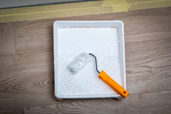 Paint roller in paint tray with white color on wooden floor.