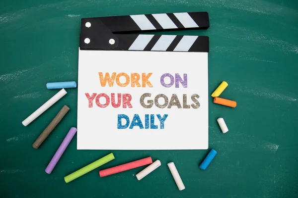 Work on your goals daily. Film clapper and colored pieces of chalk on a green chalkboard background.