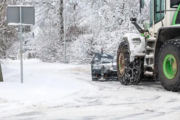 Tractor clears snow from road after snowfall, chains on tractor wheel, winter landscape.