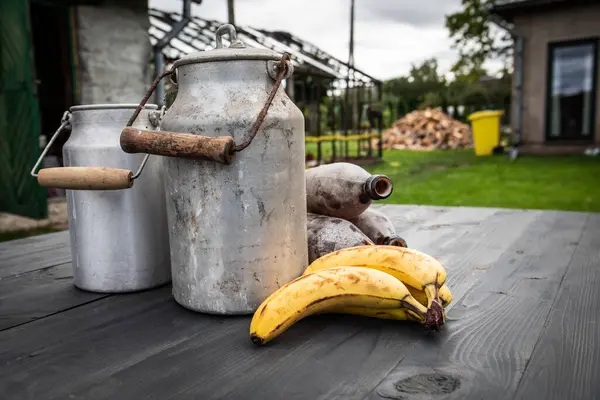 Bananas, metal cans and old glass bottles on a table in a rural setting, a yellow trash bin and a pile of firewood in the background.