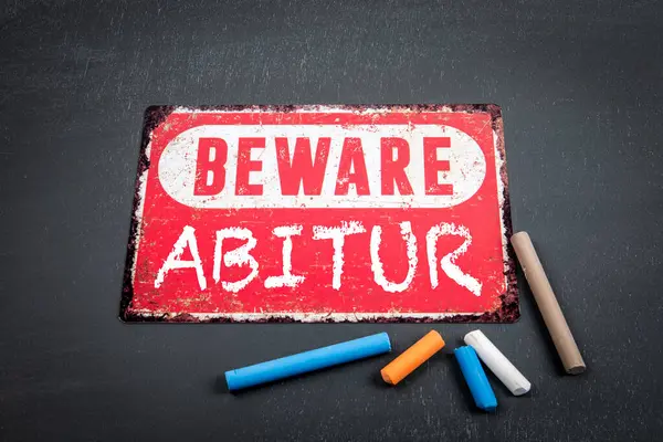 stock image High school diploma in German. Beware sign with text and colored pieces of chalk on a dark chalkboard background.