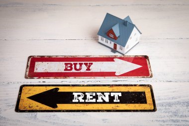 Satın al ya da Kirala. Yazılı yön okları ve beyaz ahşap desenli arka planda minyatür ev modeli..