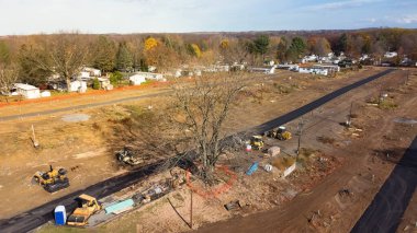 Aerial view heavy machines for foundation and earthmoving works at mobile home park construction site near row of completed manufactured trailer houses in Rochester. Upstate New York housing community