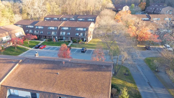 Aerial view three story barn style house apartment complex with outdoor parking and colorful fall foliage in early morning light in Penfield, New York, USA. Suburban parkside rental units