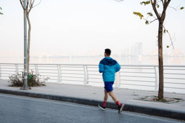Action motion blurred healthy Asian man with light jacket running along West Lake downtown buildings background early morning in Hanoi, Vietnam. Wellbeing and healthy workout at popular sightseeing