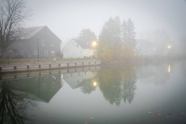 Tarihi iki katlı evler, kutup ışıkları ve Fairport Village, New York, ABD 'de renkli sonbahar nehir kenarındaki yürüyüşün yansımasıyla sisli bir sabah manzaralı Erie kanalı. Manzaralı küçük kasaba Amerika