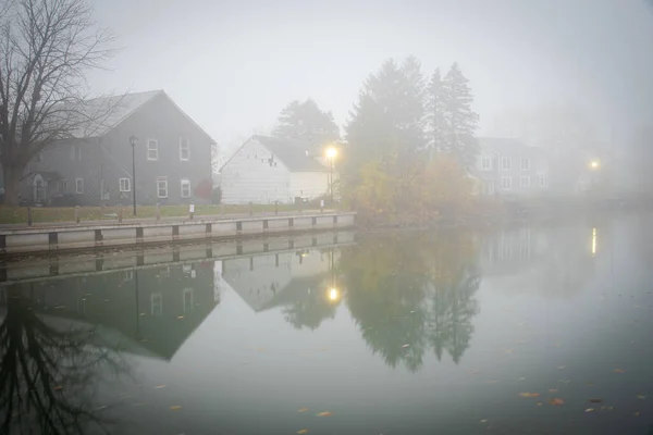Foggy morning scenic Erie canal with reflection of historic two-story houses, pole lighting and colorful fall foliage riverside walk in Fairport Village, Upstate New York, USA. Scenic small-town America