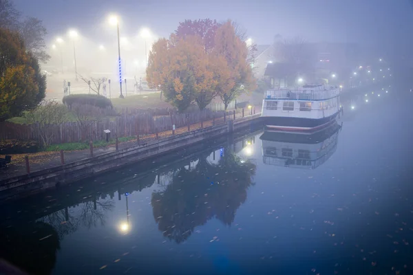 Historic Erie Canal in Fairport village with barge cruise boat tour, foggy morning mist and reflection of colorful fall foliage along riverside walk, Upstate New York, USA. Scenic small-town America