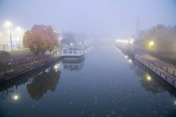Historic Erie Canal in Fairport village with barge cruise boat tour, foggy morning mist and reflection of colorful fall foliage along riverside walk, Upstate New York, USA. Scenic small-town America