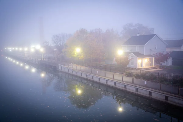 Two story house with American flag in mist foggy early morning along historic Erie Canal with yellow pole lighting, colorful fall foliage in Fairport, Upstate New York, USA. Riverside walk small town