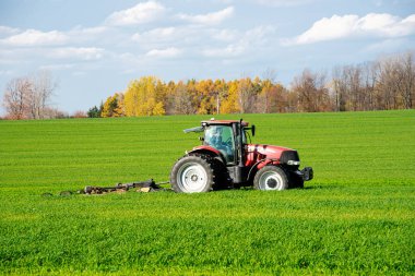 Çiftlik arazisindeki çim biçme tarlasında dönen karavanı olan daha büyük bir çiftlik traktörü, renkli sonbahar yeşillikleri, bulut mavisi gökyüzü Güney Lockport, Niagara County, Kuzey New York, ABD. Tarım endüstrisi