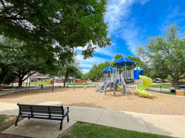 Yerel yol ve banliyö evleri yakınlarındaki büyük halk oyun parkında klasik oturma tezgahı, ahşap yoncalar üzerindeki tırmanma yapısı, uzun ağaç gölgeleri ve beton patika, Teksas, ABD. Açık hava aktiviteleri