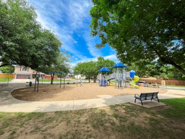 Yerel yol ve banliyö evleri yakınlarındaki büyük halk oyun parkında klasik oturma tezgahı, ahşap yoncalar üzerindeki tırmanma yapısı, uzun ağaç gölgeleri ve beton patika, Teksas, ABD. Açık hava aktiviteleri