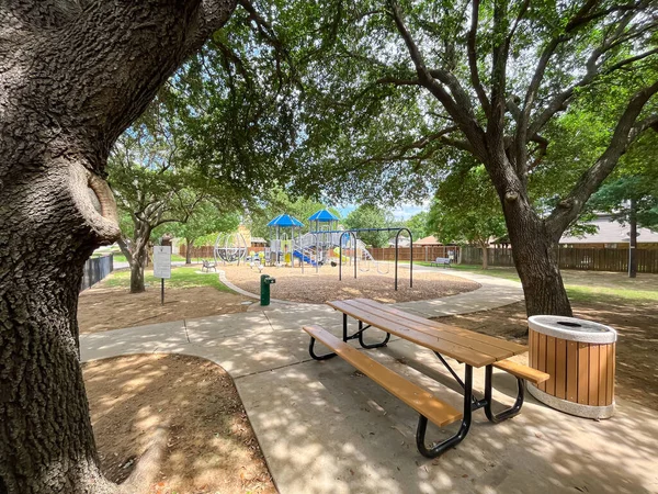 stock image Large picnic table with trash can under mature oak tree shade, drinking water fountain near playground climbing structure community park with suburban houses Grapevine, Texas, USA. Outdoor activities