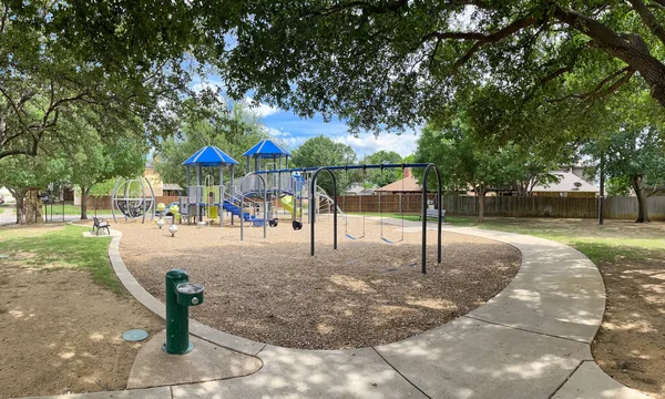 Drinking water fountain in public community park playground structure wood chips mulch, concrete sidewalk pathway, metal fence surrounding suburban houses mature trees Texas, USA. Outdoor activities