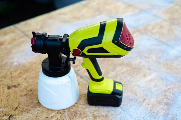 Top view cordless paint sprayer gun with full container of white paint on oriented strand board OSB board wood panel. Handy power tool for painting job.