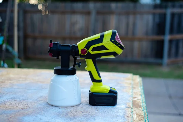 Cordless paint sprayer gun with full container of white paint on oriented strand board OSB board wood panel and blurry wooden fence background. Handy power tool for painting job