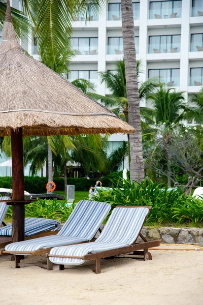 Lounge chair covered by beach umbrella thatched with palm leaves, multistory hotel background, coconut trees on white sandy shore of luxury resort in Nha Trang, Vietnam, tropical, coastline. Travel
