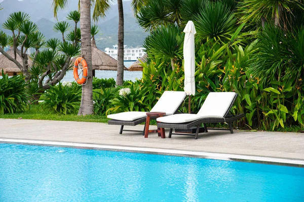 Beachside swimming pool at upscale resort with safety life buoy, lounger chairs white cushion, collapsed beach umbrella, round stool table, ocean cruise background, coconut palm tree tropical. Vietnam