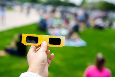 Asian hand wear smart watch holding paper solar eclipse with blurry crowd people watching totality show in Dallas, Texas, April 8, scratch resistant polymer lenses filter out harmful ultraviolet. USA clipart