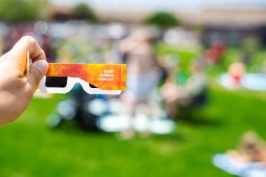 Side view of paper optics solar eclipse glasses scratch resistant polymer lenses filter harmful ultraviolet, infrared ray, blurry crow people on grassy yard watching totality show, Dallas, Texas. USA clipart