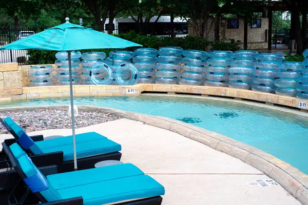 stock image Turquoise canvas outdoor chaise lounge cushion beach umbrella along lazy river full stack of clear floats transparent bathing swimming rings, 3 feet depth marker, water park hotel resort, Texas. USA