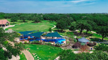 Yelken gölgesi yapısı olan ve geniş modern çocuk dinlenme merkezinde su oynayan farklı çocuklar, yatay çizgide yemyeşil ağaç, banliyö parkı, Flower Mound, Texas, hava manzaralı. ABD