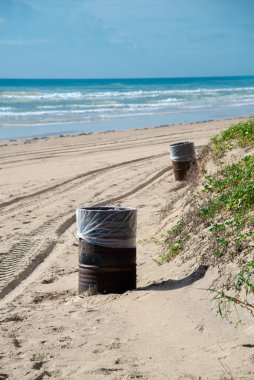 Trash can barrel with white plastic liner along white sandy shoreline with ATV tracks in South Padre Island, tropical beach of Texas, sunny blue cloud sky, littering management environmental. USA clipart