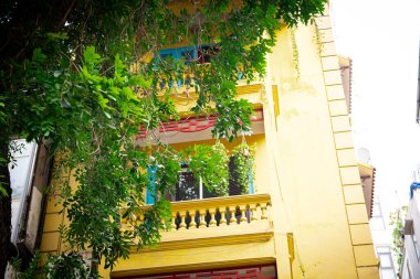 Old Quarter, Hanoi, Vietnam 'daki köşedeki ev, sarı, Fransızlardan etkilenen sömürge dönemi mimarisi, çok katlı ev, balkon ahşap panel mahremiyet korkulukları, yemyeşil. Asya