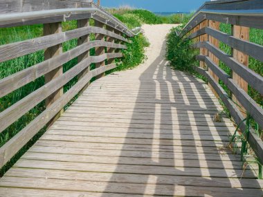 Galveston Doğu Sahili 'ne doğru giden korkuluklu tahta kaldırım ve gölge desenli yakın plan. Görüntü, dokuyu, kıyı erişimini ve güneşli gökyüzünün altındaki doğal geçişi ön plana çıkarıyor. ABD