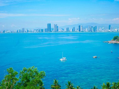 Danang parlak ufuk çizgisi okyanus ufku boyunca uzanır, sakin turkuaz suların üzerinde yükselir. Ön planda, bereketli tropikal ağaçlar ve palmiyeler şehir kıyı manzarasına canlı bir kontrast ekler. Vietnam