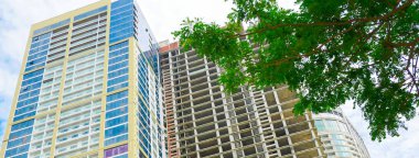 Panorama manzarası alçak açılı, yeşil yapraklarla çerçevelenmiş iskelet beton yaprağının yanında cam kuleyi gösteriyor, Danang 'da gelişen ufuk çizgisinin doğal ve kentsel gelişimi arasındaki zıtlığı yakalıyor. Vietnam