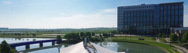 Panorama aerial view pavilion amphitheater beside North Lake features tensile roof, pedestrian bridge, trails, glass office tower, ideal for lakeside events and urban leisure in Cypress Water, TX. USA