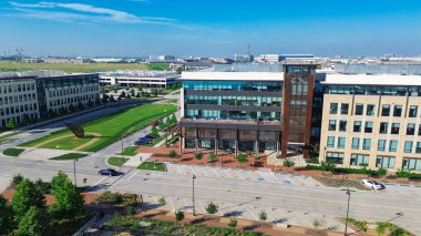 Street-level view of glass-walled office building with patio seating along Cypress Waters Boulevard near DFW corridor, multilevel parking garage with canopy, architectural sculpture, vacant land. USA