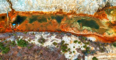 This aerial photograph shows the devastating impact of industrial processes on the environment. A heavily polluted river is seen with toxic green waste and orange deposits on the banks, while the adjacent vegetation has been severely affected and sup