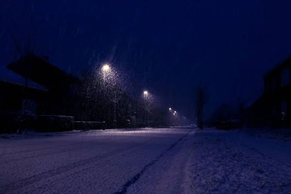 A wide dark night portrait of a snowy street during heavy snowfall with street lights illuminating the way for cars. The road looks very slippy and should be cleared, because it is dangerous.
