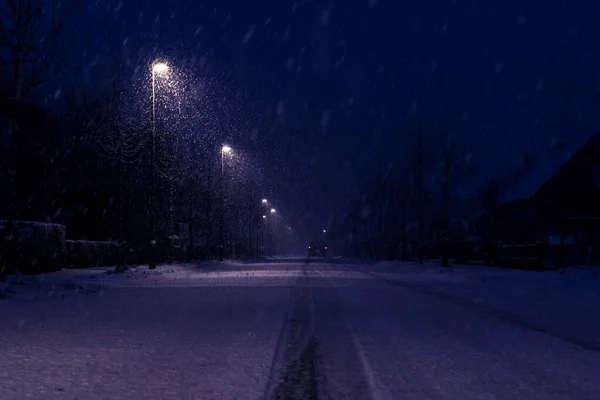 A dark night portrait of a snowy street with snow flakes still falling with street lights illuminating the way for cars. The road looks very slippy and should be cleared, because it is dangerous.