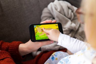 Brecht, Belçika - 23 Ekim 2022: Anne akıllı telefonu elinde tutarken bir çocuğun mobil oyun uygulamasını oynaması portresi. Küçük çocuk ekranı gösteriyor ve sürüklüyor..