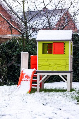 Küçük yeşil bir oyun evinin portresi, kışın karla kaplı bir bahçede duran kırmızı bir kaydırak. Evin küçük kırmızı pencereleri ve bir kapısı var ve oynanmaya hazır..