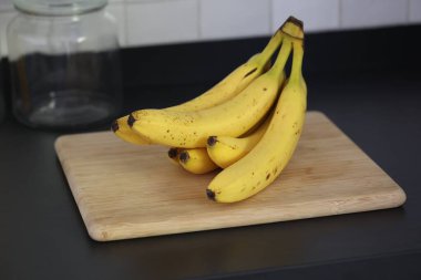 Siyah bir mutfak tezgahının tahta kesme tahtasının üzerinde duran bir grup sarı muzun portresi. Lezzetli enerji veren yemekler yemeğe hazır ve spordan önce ideal..