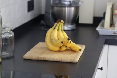 Siyah bir mutfak tezgahının tahta kesme tahtasının üzerinde duran bir grup sarı muzun portresi. Lezzetli enerji veren yemekler yemeğe hazır ve spordan önce ideal..