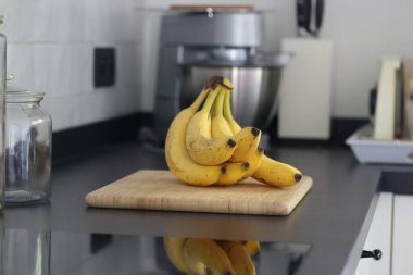 Siyah bir mutfak tezgahının tahta kesme tahtasının üzerinde duran bir grup sarı muzun portresi. Lezzetli enerji veren yemekler yemeğe hazır ve spordan önce ideal..