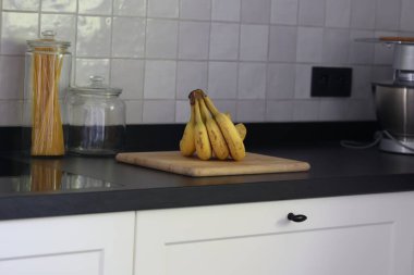 Siyah bir mutfak tezgahının tahta kesme tahtasının üzerinde duran bir grup sarı muzun portresi. Lezzetli enerji veren yemekler yemeğe hazır ve spordan önce ideal..