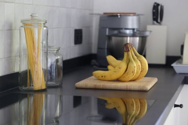 Siyah bir mutfak tezgahının tahta kesme tahtasının üzerinde duran bir grup sarı muzun portresi. Lezzetli enerji veren yemekler yemeğe hazır ve spordan önce ideal..